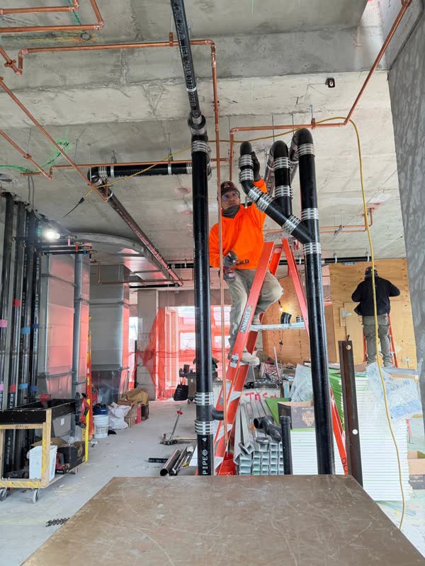 Plumber on a ladder connecting angled cast-iron vent branches to a vertical riser, overhead copper mains behind