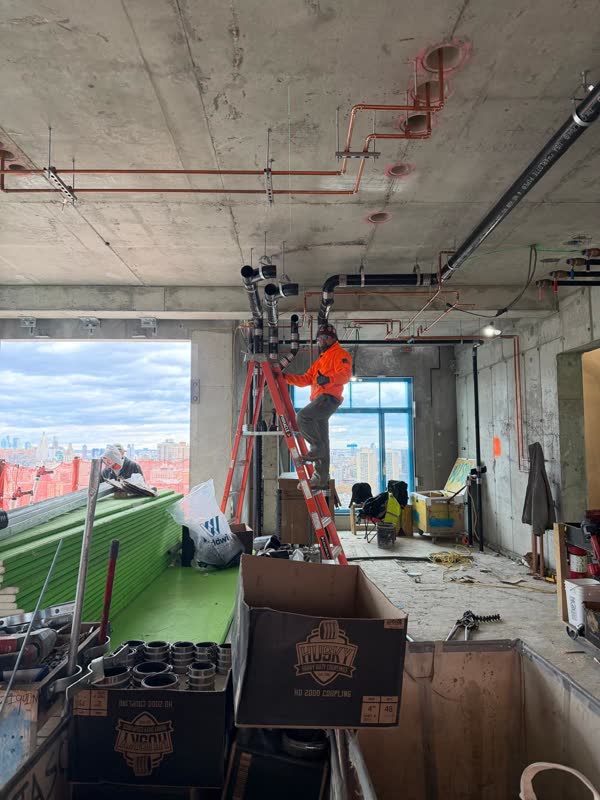 Copper water mains strapped to a concrete deck beside cast-iron vent takeoffs, plumber on a ladder, city skyline through…