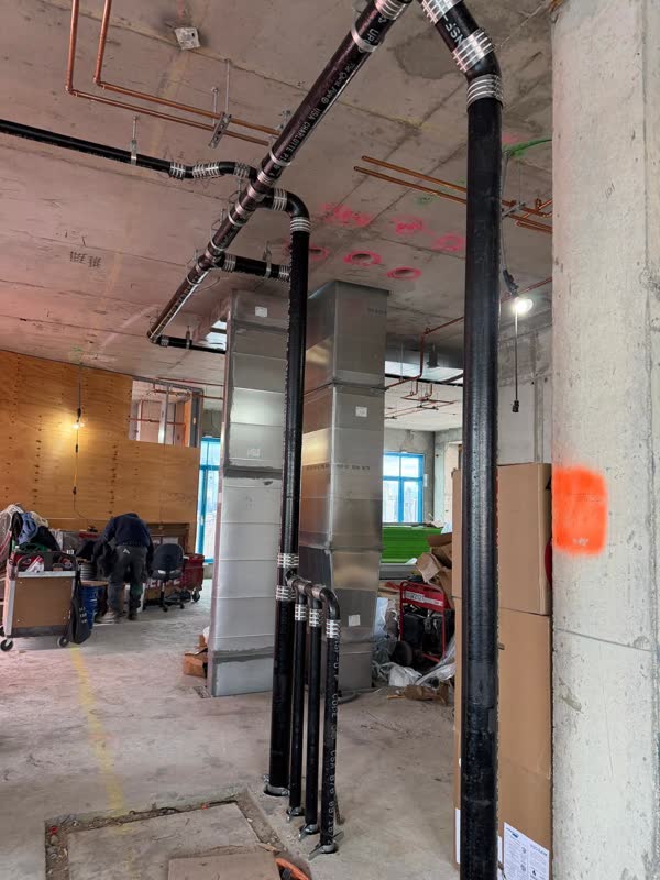 Bank of cast-iron no-hub risers turning into an overhead waste line next to sheet-metal duct on a concrete floor