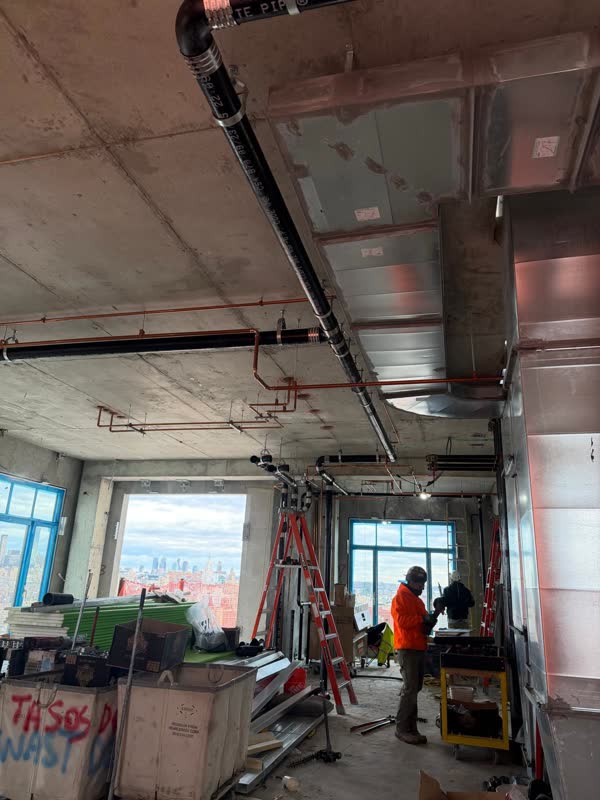 Overhead copper water mains and a sloped cast-iron waste line routed alongside sheet-metal duct on a high-rise floor