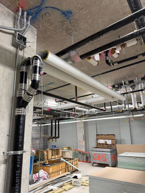 Insulated white domestic water mains and black steel piping hung in tidy rows beside a 4-inch cast-iron no-hub stack