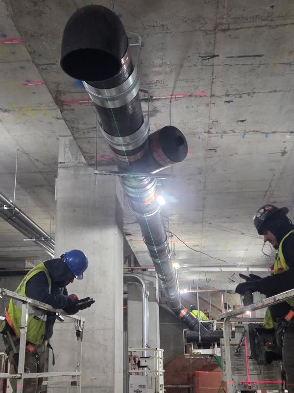 Two plumbers on scissor lifts joining a large-diameter cast-iron riser and branch to a concrete deck with laser layout line