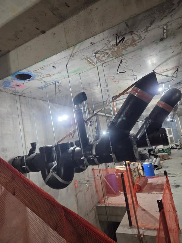 Cast-iron no-hub waste manifold with wye branches and shielded couplings hung on threaded rod below a concrete deck