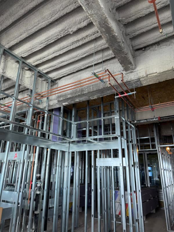 Overhead run of copper water mains on trapeze hangers under a fireproofed concrete deck above steel-stud partition framing