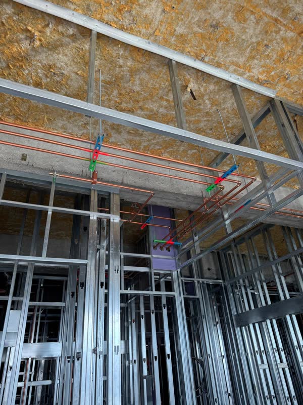 Parallel copper hot and cold mains banked on color-coded pipe hangers below deck, above steel-stud framing