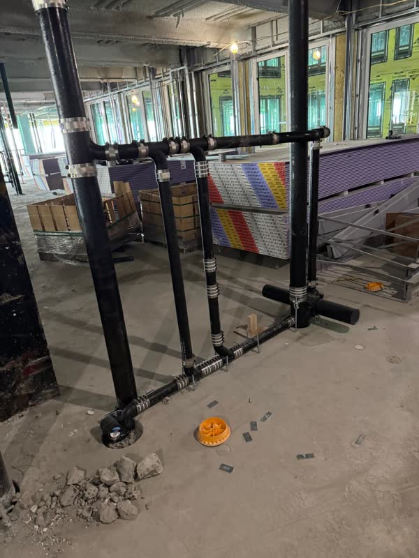 Cast-iron no-hub waste header with four risers, strapped to the slab beside stacked drywall on a fitted-out floor