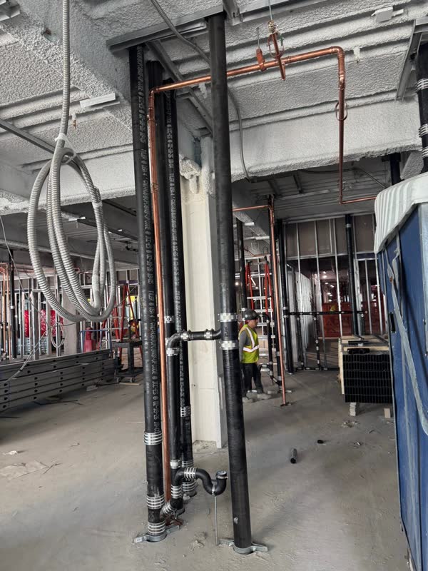 Cast-iron risers beside a fireproofed column with overhead copper main and a hanger drop on a commercial floor