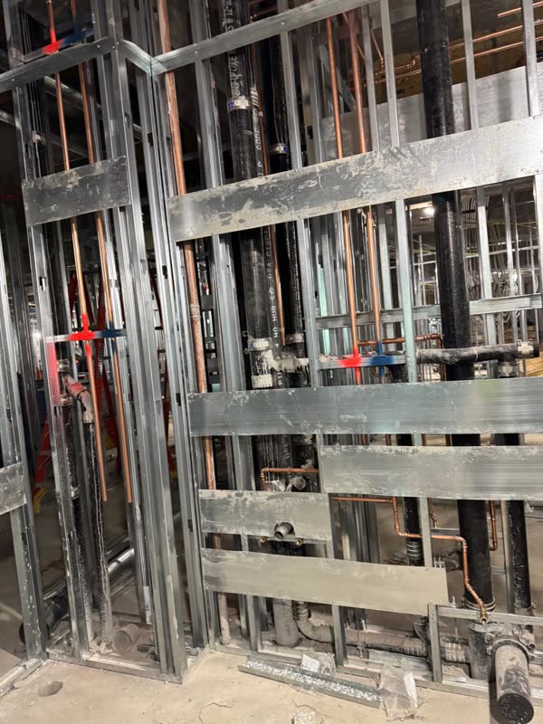 Copper supply lines on color-coded clips and cast-iron no-hub stacks running inside steel-stud walls with backing plates