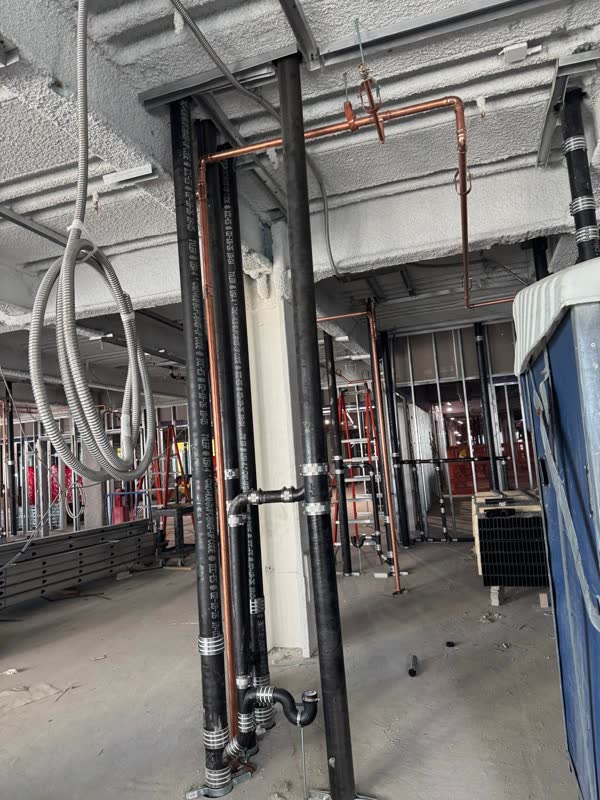 Cast-iron no-hub waste risers beside a copper water main branching along a fireproofed concrete deck