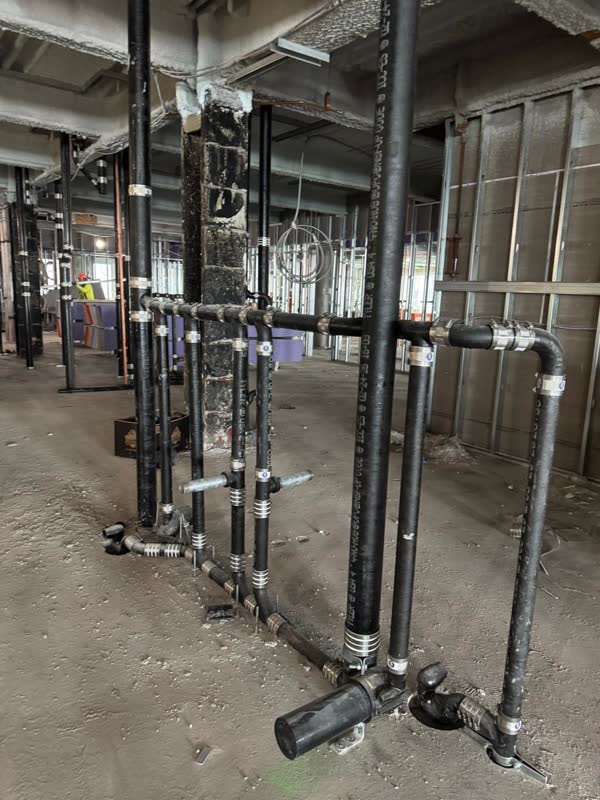 Row of cast-iron no-hub risers tied into a horizontal waste header with no-hub bands along a concrete floor