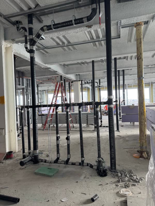 Overhead cast-iron waste main sweeping into a battery of no-hub floor risers on a bright high-rise floor