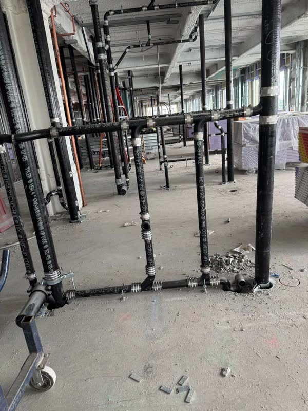 Grid of cast-iron no-hub drain lines and risers laid across a concrete floor with copper water lines behind