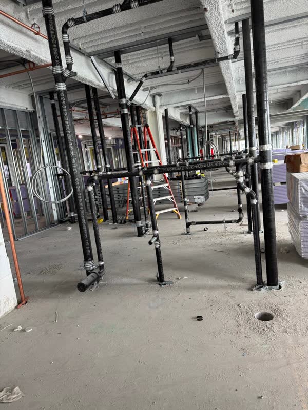 Overhead cast-iron no-hub waste mains and a bank of risers braced to the slab across an open commercial floor
